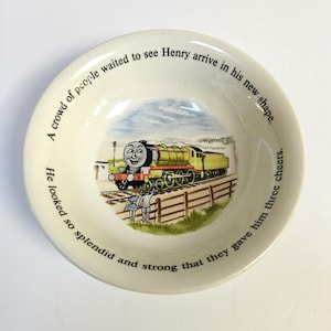 May include: Cream-colored ceramic bowl featuring a colorful illustration of a green and yellow train with a smiling face. The bowl has text around the rim that reads, "A crowd of people waited to see Henry arrive in his new shape."