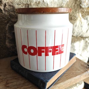 May include: White ceramic coffee canister with a wooden lid. The canister has red vertical stripes and the word "COFFEE" in red block letters. The canister is sitting on a blue book.