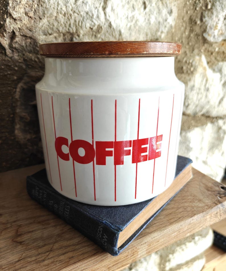 May include: White ceramic coffee canister with a wooden lid. The canister has red vertical stripes and the word "COFFEE" in red block letters. The canister is sitting on a blue book.
