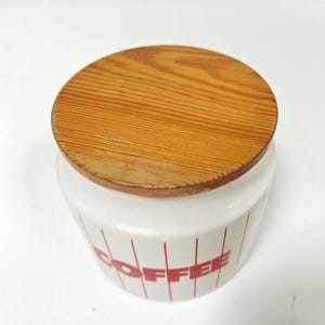May include: A white ceramic coffee canister with a wooden lid. The canister has red vertical stripes and the word "COFFEE" in red. The lid is round and made of light brown wood.