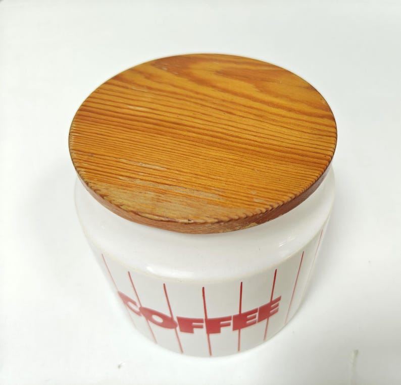 May include: A white ceramic coffee canister with a wooden lid. The canister has red vertical stripes and the word "COFFEE" in red. The lid is round and made of light brown wood.