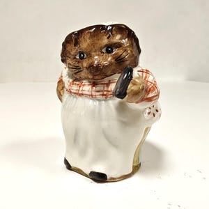 以下が含まれることがあります： 白いエプロンと赤と白のチェック柄のスカーフを着た、茶色の動物（おそらくアナグマまたは同様の生き物）の陶器製の置物。置物は黒い物体を持っており、黒い塗装が施されています。