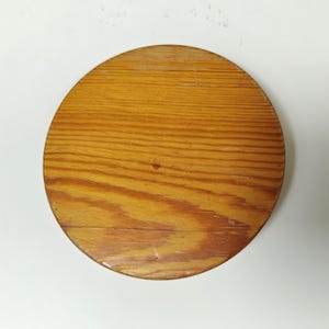 May include: A round wooden object with a light brown finish and visible wood grain. The object appears to be a lid or a coaster, with a diameter of approximately 8 inches.