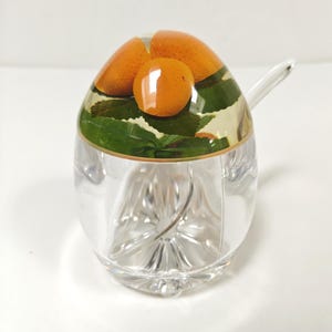 May include: A clear, decorative sugar bowl with a lid featuring orange fruit and green leaves. The bowl has a spoon and a faceted design on the lower half. The lid has a gold trim.