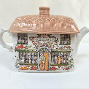 Peut inclure: Théière en céramique en forme de cottage, avec un toit de chaume et une façade représentant un magasin avec le texte "Ye Olde Tea Pot Inn". La théière est décorée de détails floraux et architecturaux, et possède une anse et un bec verseur.