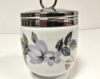 Pode incluir: Ovo de cerâmica branca com tampa e alça prateadas. O ovo é decorado com um padrão floral em tons de roxo, cinza e rosa. O ovo tem uma base.