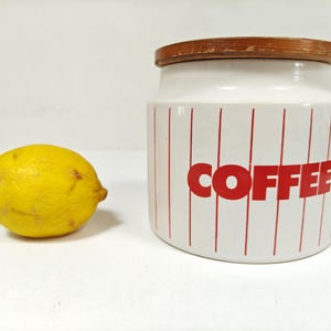 May include: A white ceramic coffee canister with red vertical stripes and the word "COFFEE" in red block letters. A yellow lemon sits to the left of the canister. The canister has a wooden lid.