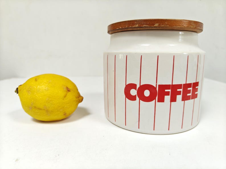 May include: A white ceramic coffee canister with red vertical stripes and the word "COFFEE" in red block letters. A yellow lemon sits to the left of the canister. The canister has a wooden lid.