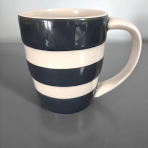 May include: A ceramic mug with a cream-colored interior and handle. The exterior features alternating horizontal bands of navy blue and cream. The mug is designed with a curved shape and a large handle for easy gripping.