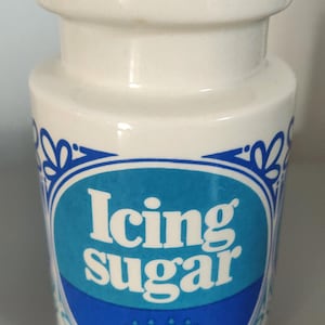 Puede incluir: Tarro de cerámica blanco con la inscripción "Icing Sugar" en un diseño azul y blanco. El tarro presenta un óvalo azul con el texto y una ilustración de pastel. El tarro tiene tapa y está diseñado para uso en la cocina.