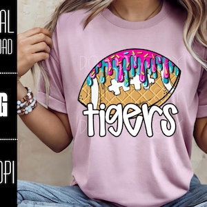 May include: A light pink t-shirt featuring a football design with a waffle cone texture, topped with pink and blue drizzles. The word "tigers" is written in white, outlined in black. The shirt is worn by a person.