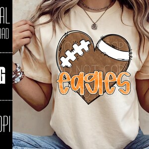 Puede incluir: Camiseta de color beige claro con un dise&ntilde;o de bal&oacute;n de f&uacute;tbol en forma de coraz&oacute;n y la palabra "Eagles" en naranja. El bal&oacute;n de f&uacute;tbol es marr&oacute;n con detalles blancos. Las palabras "DIGITAL DOWNLOAD", "PNG" y "300 DPI" est&aacute;n en el lado izquierdo.