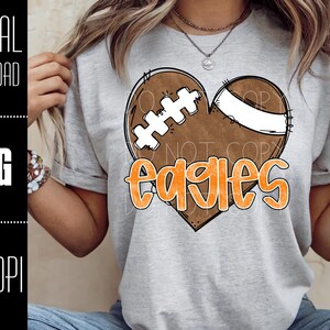 Puede incluir: Camiseta gris claro con un dise&ntilde;o de bal&oacute;n de f&uacute;tbol en forma de coraz&oacute;n y la palabra "Eagles" en naranja. El bal&oacute;n es marr&oacute;n con cordones blancos y una franja blanca. A la izquierda, el texto dice "DIGITAL DOWNLOAD", "PNG" y "300 DPI".