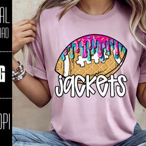May include: A light pink t-shirt featuring a football design with dripping pink and blue icing. The word "jackets" is written in a playful font below the football graphic. The image is a digital download.
