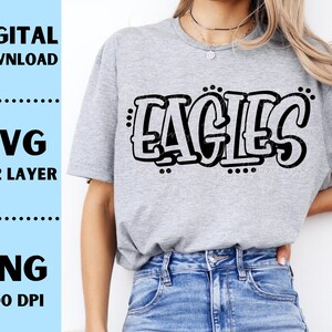 Op de afbeelding: Lichtgrijs t-shirt met het woord "EAGLES" in een zwarte, omlijnde letter. De tekst is in graffiti-stijl. De afbeelding bevat ook de woorden "DIGITALE DOWNLOAD", "SVG 1 & 2 LAYER" en "PNG 300 DPI" in een lichtblauwe box.