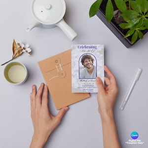 May include: A memorial card with a photo of a smiling person, framed in a blue arch, with the text "Celebrating His Life". The card is held near a brown envelope, a white pen, and a cup of tea.