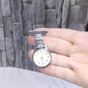 May include: A silver nurse's fob watch with a skateboard-shaped clip. The watch has a white face with black numerals and red hands. The watch is attached to a silver metal band.