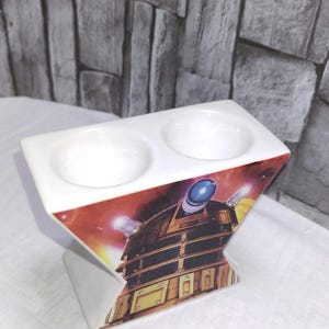 May include: White ceramic double egg cup featuring a Dalek design from Doctor Who. The egg cups are circular and recessed. The base of the egg cup has a triangular shape with a red and orange background.