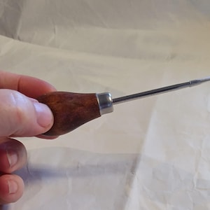 May include: A scratch awl featuring a smooth, dark brown wooden handle with a rounded, ergonomic shape and a silver-coloured metal ferrule connecting to a long, slender, pointed steel shaft with a protective clear plastic tip cover.