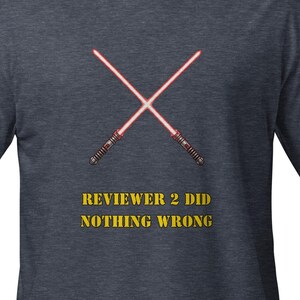 Può includere: T-shirt blu scuro con due spade laser rosse incrociate. Il testo "REVIEWER 2 DID NOTHING WRONG" è stampato in giallo sotto. La maglietta è a girocollo.