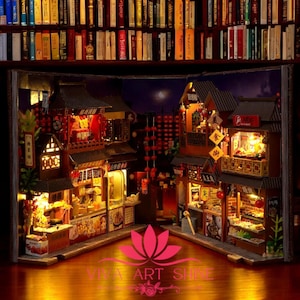 May include: A miniature wooden model of a Chinese street scene with red lanterns, food stalls, and buildings. The model is lit up with small lights and is open like a book.