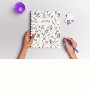May include: A white weekly planner with a floral design featuring green leaves and white flowers. The planner has a black spiral binding and the text "WEEKLY PLANNER" and "Every Moment Matters".