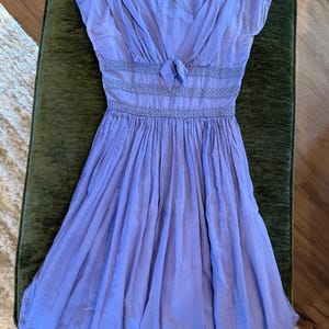 May include: A light purple midi dress featuring short sleeves, a V-neckline with a center tie detail, and two horizontal bands of decorative smocking at the waist. The skirt is gathered and flows downward from the smocked waistline.