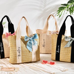 May include: Four straw tote bags with personalized names on the front. Each bag has a different colored handle and a matching ribbon or scarf tied to the handle. The bags are sitting on a white surface.