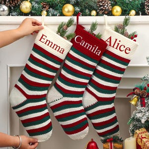 May include: Three personalized Christmas stockings with red, green, and white stripes are displayed. The stockings are labeled "Emma", "Camila", and "Alice" in red text. The stockings are hanging on a white mantelpiece decorated with greenery and ornaments.