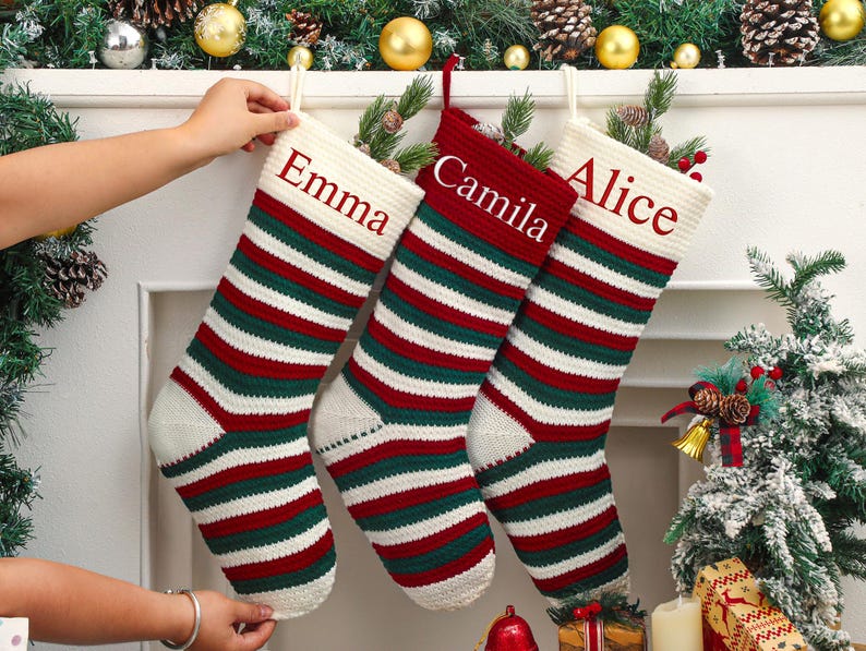 May include: Three personalized Christmas stockings with red, green, and white stripes are displayed. The stockings are labeled "Emma", "Camila", and "Alice" in red text. The stockings are hanging on a white mantelpiece decorated with greenery and ornaments.