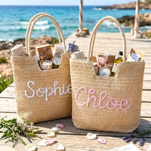 May include: Two woven straw tote bags with two top handles each. One bag features the name Sophie embroidered in white script, and the other features the name Chloe embroidered in pink script. Both bags are filled with various gift items and small boxes.