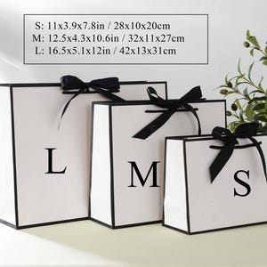 May include: Three white gift bags with black ribbon bows and black trim. The bags are labelled with the letters "S", "M", and "L". The bags are on a white surface. The text on the image reads: "S: 11x3.9x7.8in / 28x10x20cm M: 12.5x4.3x10.6in/32x11x27cm L: 16.5x5.1x12in/42x13x31cm"