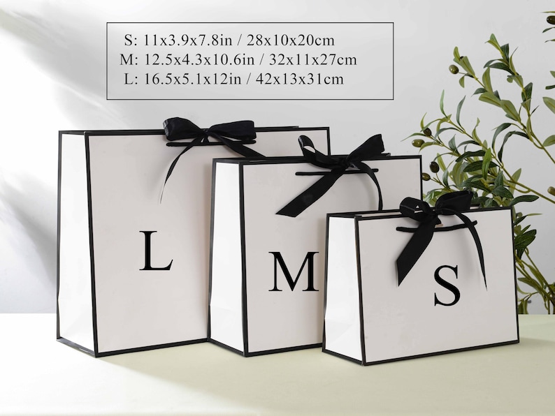 May include: Three white gift bags with black ribbon bows and black trim. The bags are labelled with the letters "S", "M", and "L". The bags are on a white surface. The text on the image reads: "S: 11x3.9x7.8in / 28x10x20cm M: 12.5x4.3x10.6in/32x11x27cm L: 16.5x5.1x12in/42x13x31cm"