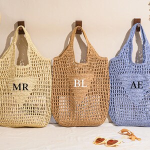May include: Three open-weave tote bags in beige, tan, and light blue, each with a heart-shaped pocket. The beige bag has "MR" printed on it, the tan bag has "BL", and the blue bag has "AE". A straw hat and sunglasses are also visible.