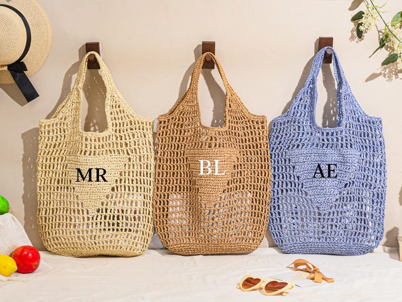 May include: Three open-weave tote bags in beige, tan, and light blue, each with a heart-shaped pocket. The beige bag has "MR" printed on it, the tan bag has "BL", and the blue bag has "AE". A straw hat and sunglasses are also visible.