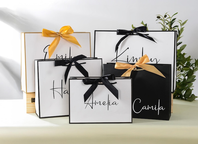 May include: Five white gift bags with black trim and black ribbon bows. The bags are labelled with the names Amelia, Hailey, Emily, Kinsley, and Camila.