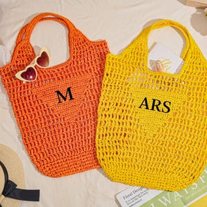 May include: Two open-weave crochet tote bags, one orange and one yellow, with large handles. The orange bag has the letter "M" and heart-shaped sunglasses. The yellow bag has the letters "ARS".