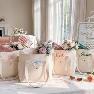 Pode incluir: Quatro sacos de lona em tom cru com bolsos exteriores e fechos de pressão. Cada saco apresenta bordados personalizados em cores diferentes com os nomes M, Taylor, Hailey e Jessica. Os sacos estão preenchidos com arranjos florais.