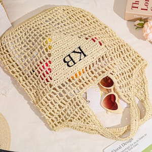 May include: A beige woven tote bag with the initials "KB" in black. The bag contains sunglasses, a book, and colorful items. A straw hat and flowers are also visible.