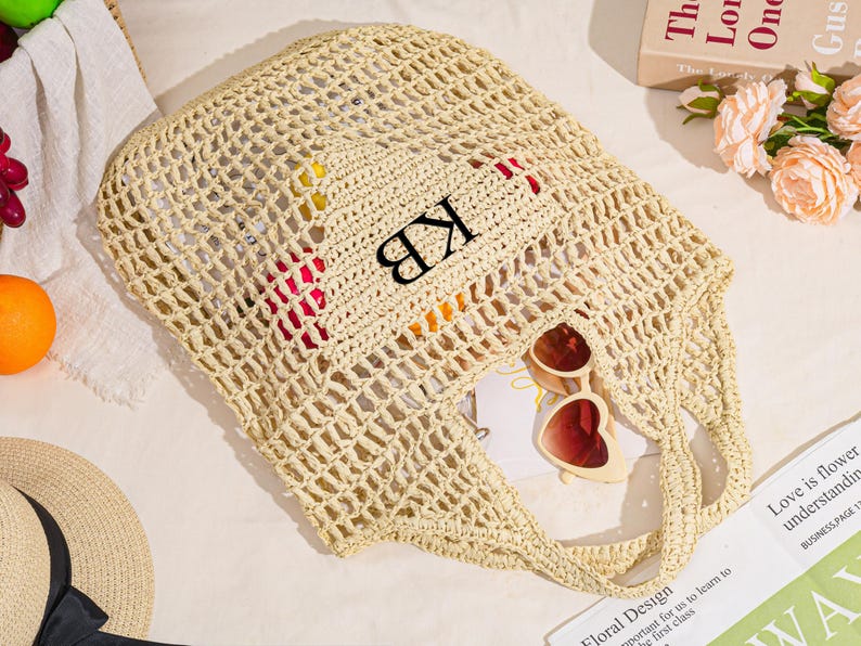 May include: A beige woven tote bag with the initials "KB" in black. The bag contains sunglasses, a book, and colorful items. A straw hat and flowers are also visible.