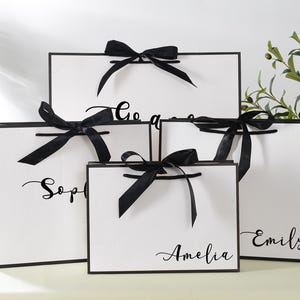 May include: Five white gift bags with black handles and black ribbon bows. The bags are stacked on top of each other. The bags are personalised with the names "Sophia", "Amelia", "Emily", and "Go Away".