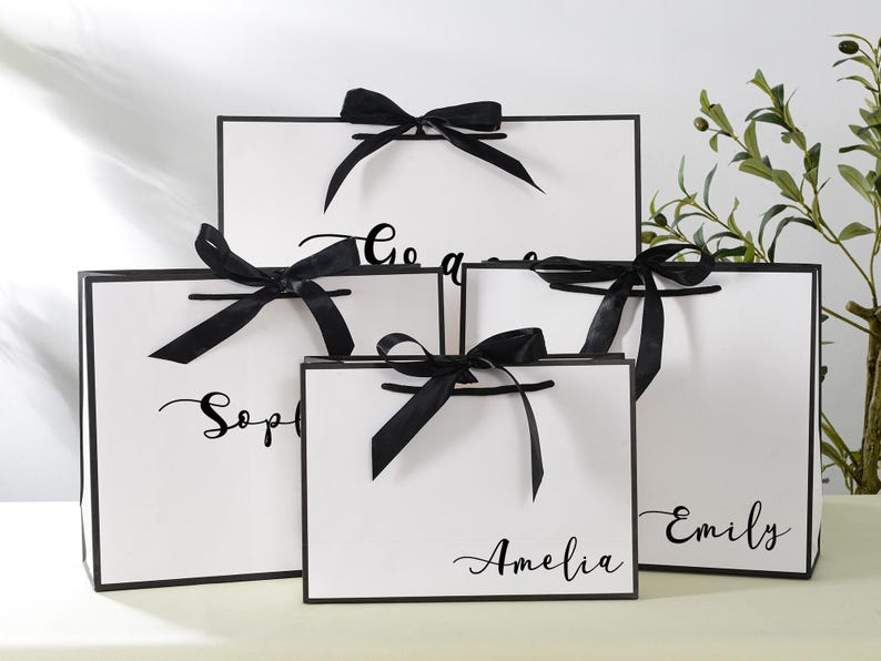 May include: Five white gift bags with black handles and black ribbon bows. The bags are stacked on top of each other. The bags are personalised with the names "Sophia", "Amelia", "Emily", and "Go Away".