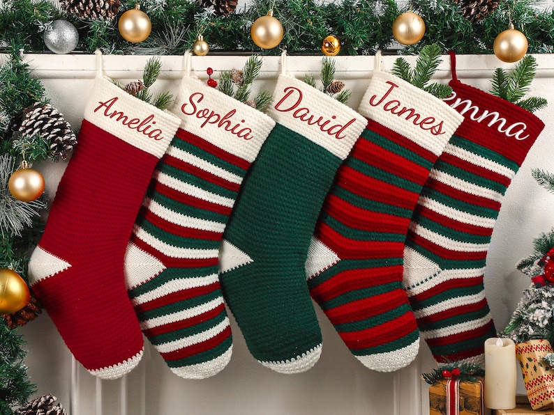 May include: Six personalized Christmas stockings in red, green, and white, each with a name embroidered in red. The stockings are hanging from a white mantelpiece decorated with greenery, pinecones, and gold ornaments.