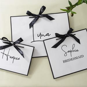 May include: Three white gift bags with black ribbon bows and black calligraphy writing. The bags say "Harper", "Luna", and "Sophia Bridesmaid".