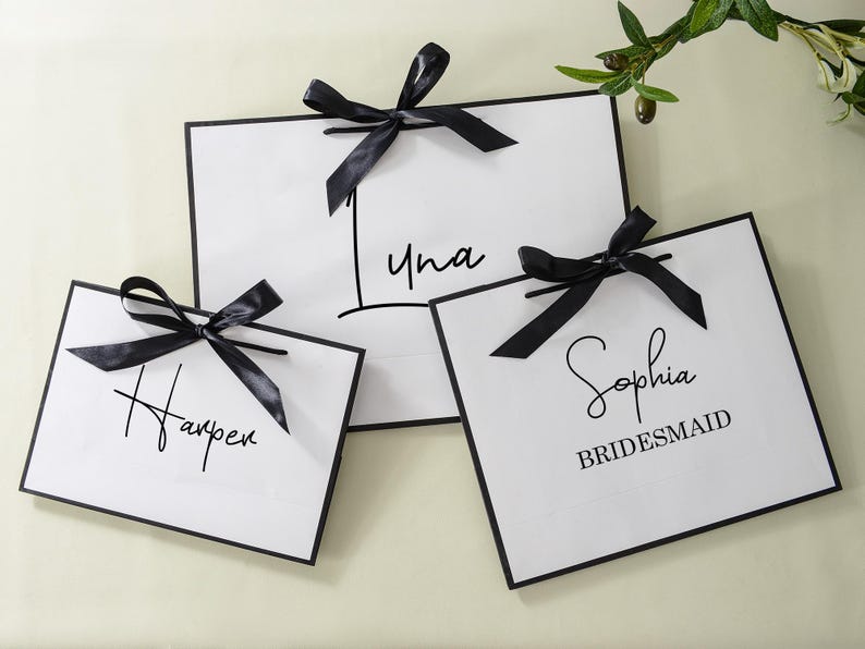 May include: Three white gift bags with black ribbon bows and black calligraphy writing. The bags say "Harper", "Luna", and "Sophia Bridesmaid".