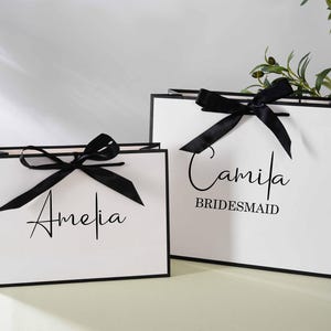 May include: Two white gift bags with black ribbon bows and black trim. The bag on the left has the name "Amelia" written on it. The bag on the right has the name "Camila" written on it, along with the word "Bridesmaid".