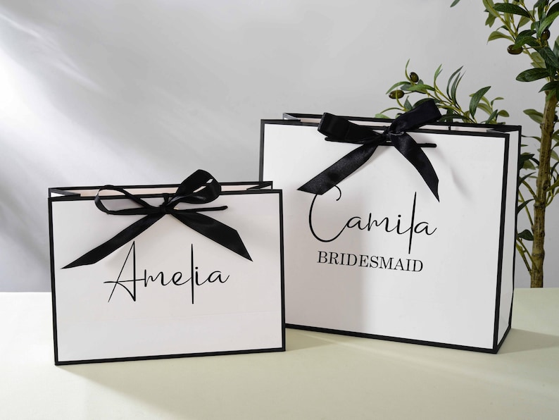 May include: Two white gift bags with black ribbon bows and black trim. The bag on the left has the name "Amelia" written on it. The bag on the right has the name "Camila" written on it, along with the word "Bridesmaid".