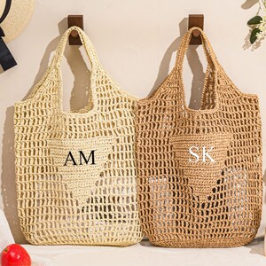 May include: Two woven straw tote bags, one light beige and one brown, hanging on wooden hooks. The beige bag has the initials "AM" and the brown bag has "SK". Both bags have a heart-shaped design.