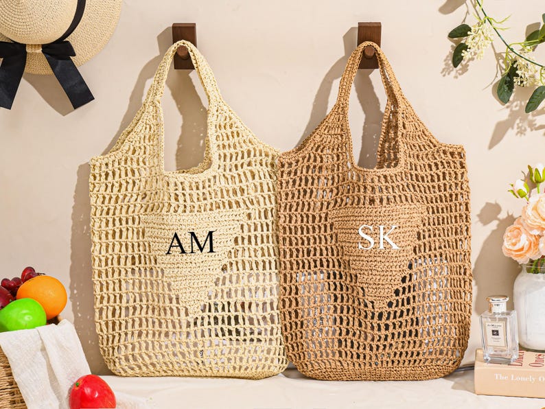 May include: Two woven straw tote bags, one light beige and one brown, hanging on wooden hooks. The beige bag has the initials "AM" and the brown bag has "SK". Both bags have a heart-shaped design.