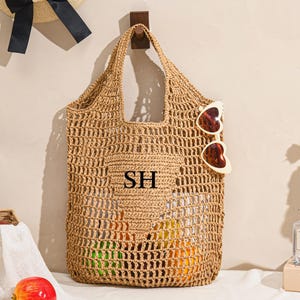 May include: A woven straw tote bag with a large open weave design. The bag has a pocket with the initials "SH" and is hanging on a wooden hook. Heart-shaped sunglasses are attached to the bag. A straw hat with a black bow hangs above.